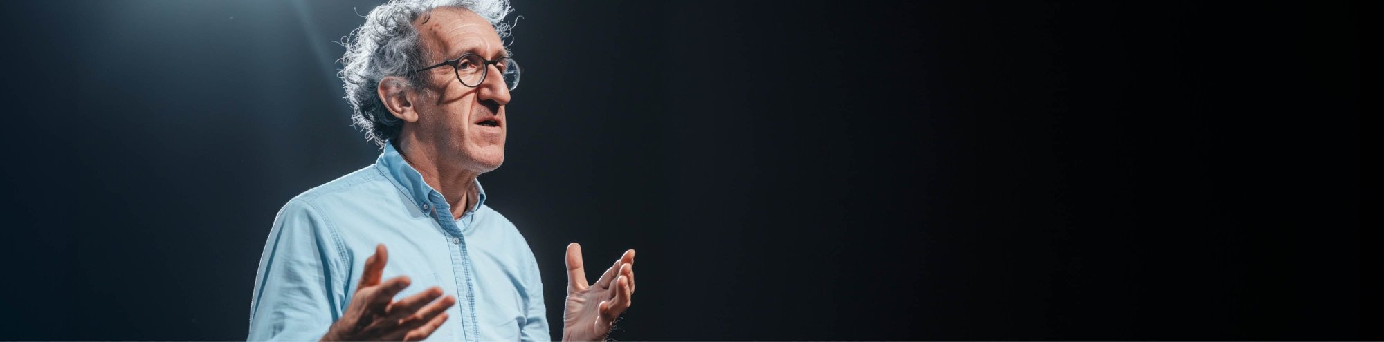 Älterer Mann mit Brille und grauem Haar spricht vor dunklem Hintergrund. Er trägt ein hellblaues Hemd und gestikuliert mit den Händen, als würde er etwas erklären oder vortragen.