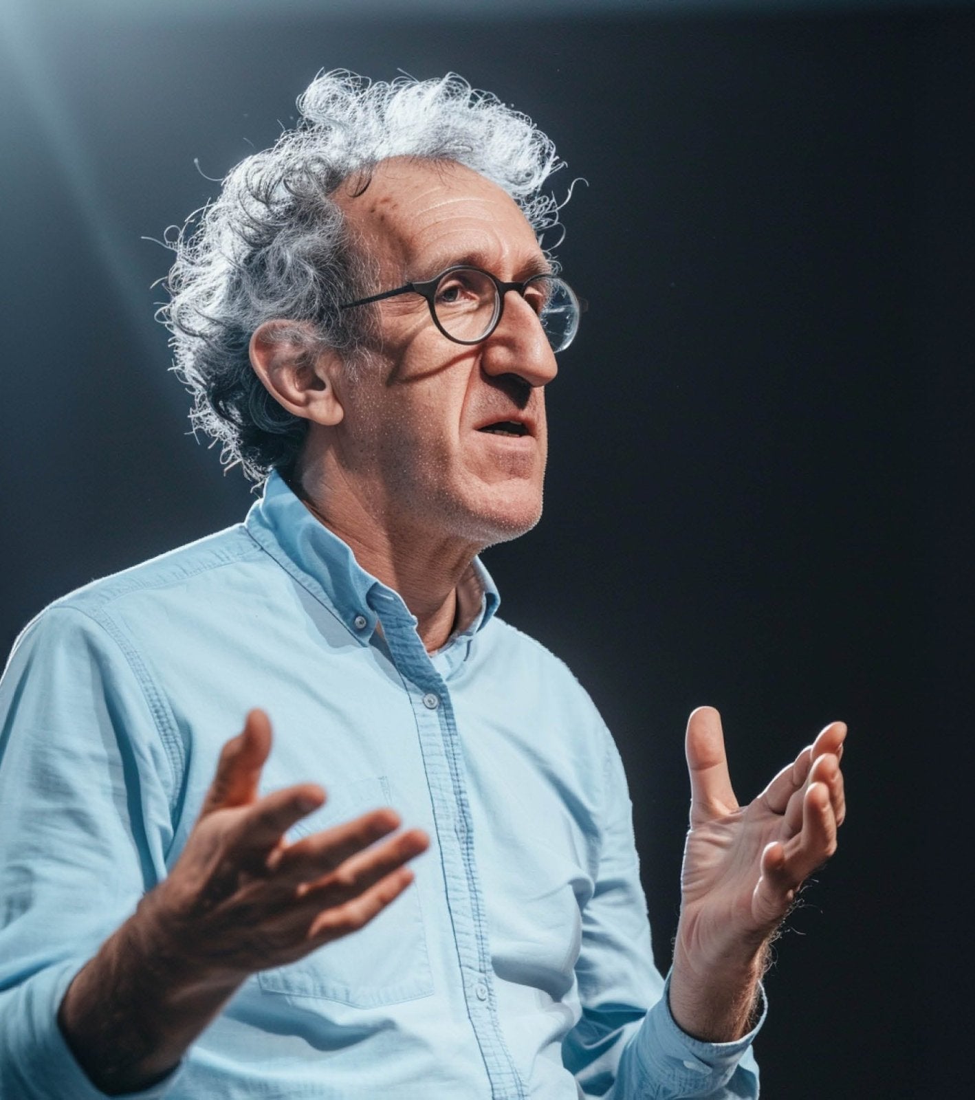 Älterer Mann mit Brille und grauem Haar spricht vor dunklem Hintergrund. Er trägt ein hellblaues Hemd und gestikuliert mit den Händen, als würde er etwas erklären oder vortragen.