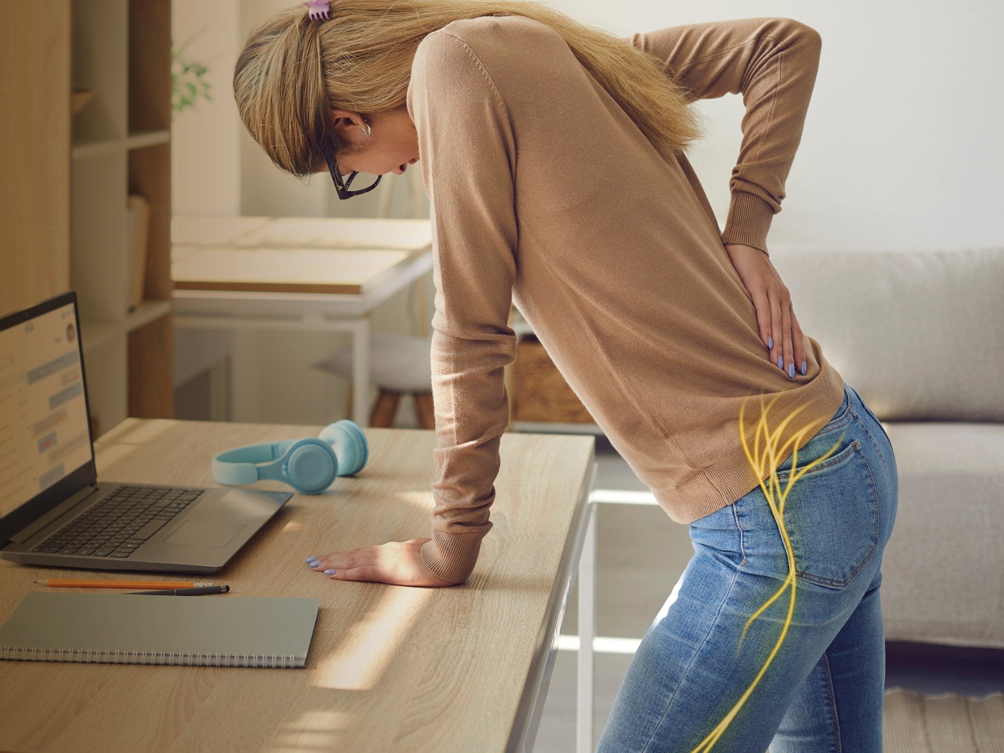 Junge Frau stützt sich auf einen Tisch und greift sich an den unteren Rücken. Eine gelbe Nervenzeichnung auf ihrer Jeans visualisiert Ischias- oder Rückenschmerzen.
